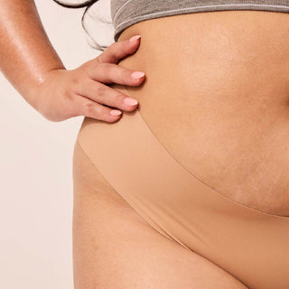 Close up image of a woman with her hand on her hip, she has a curvier figure and glowing, dewy skin. She is wearing light tan coloured underwear.