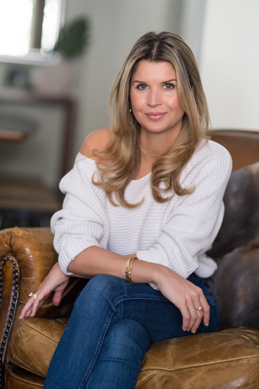 Photo of Lexi Miles Corrin, owner and founder of WAXON, sat on a couch, wearing a white sweater and blue jeans. 