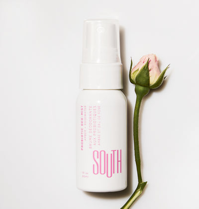 Top down view of SOUTH Deomist bottle, a PH Balanced spray for intimate parts of the body. A white bottle with pink writing laid next to a rose.
