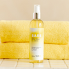 BARE body oil bottle, shot in front of a stack of yellow towels on a tiled surface.