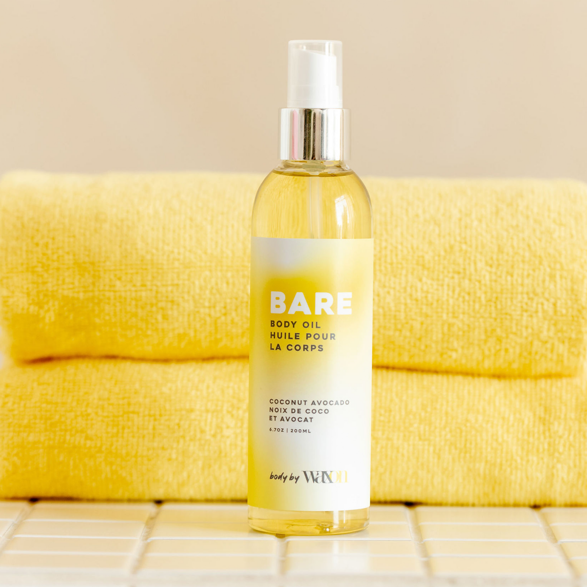 BARE body oil bottle, shot in front of a stack of yellow towels on a tiled surface.
