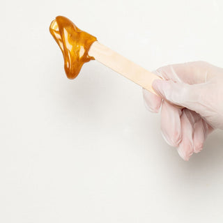 A gloved hand holding a wax stick with warm, dripping gold hair removal wax on the end. The gold wax is WAXON's proprietary hair removal wax formula.