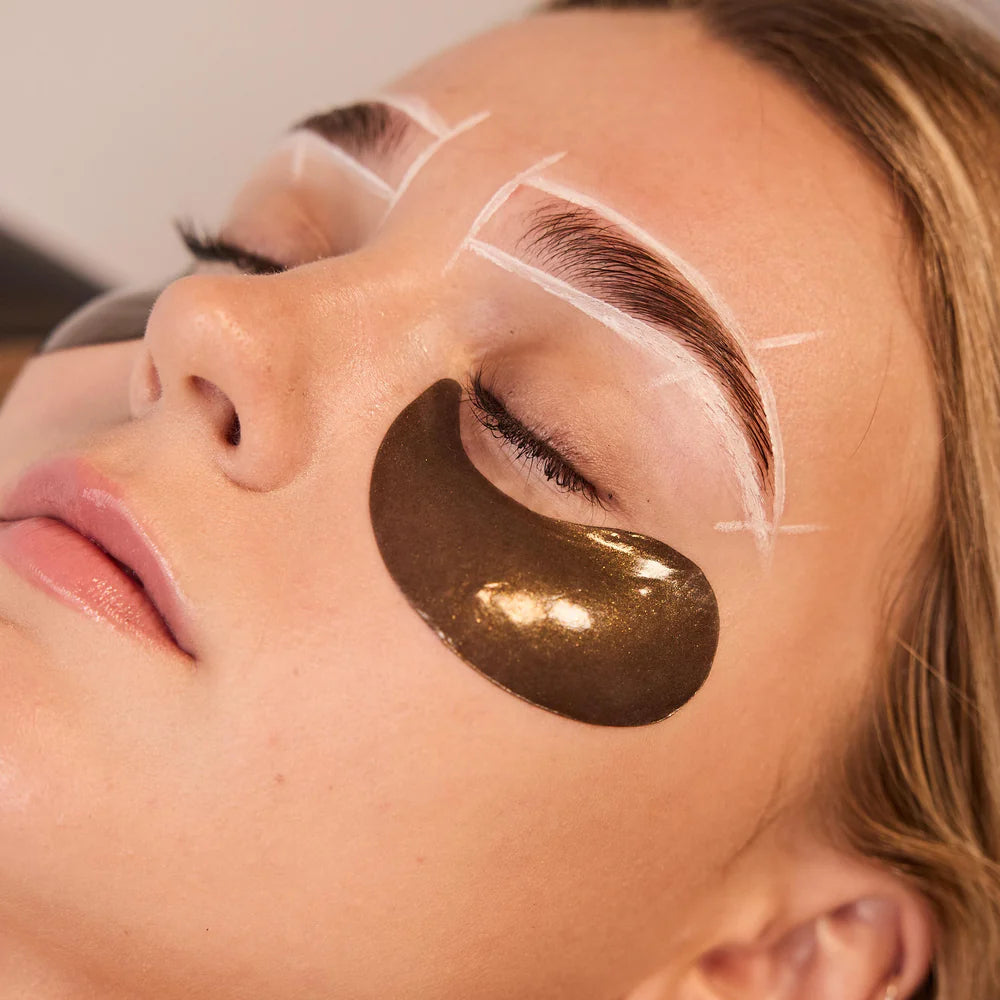 An image of a woman receiving WAXON's Brow Boost all-inclusive brow service. She has brow mapping on her brows and Radford Gold Eye patches on during the service.