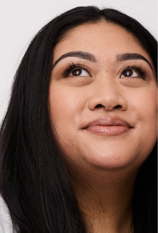 Portrait of a young woman looking up to the left of the image, she has a slight smile and glowy skin. Her freshly shaped, tinted, and waxed eyebrows are the focus of the image